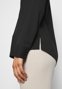 Vue latérale d'une personne portant une chemise noire à manches longues avec des poignets boutonnés et un pantalon beige clair sur un fond uni.