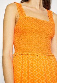 Femme portant une robe orange sans manches avec un corsage smocké texturé et une jupe à motifs géométriques.
