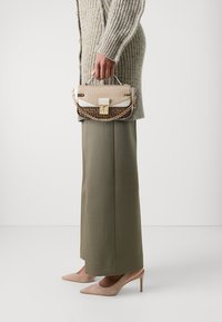 Beige handbag with geometric pattern, gold hardware, and a chain strap, held next to a textured knit sweater and olive green wide-leg pants.