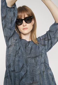 Blusa azul com padrão de linhas onduladas, material leve e mangas volumosas. Óculos de sol pretos com armação angular.