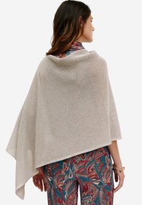 Vaaleanbeige neulottu poncho, jossa on avoin kudontamalli, valuvat olat ja sileä tekstuuri, yhdistettynä värikkäisiin kuviollisiin housuihin.