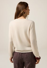 Suéter de punto crema con mangas largas y cuello redondeado, combinado con pantalones burdeos texturizados. Los detalles incluyen puños y cinturilla acanalados.