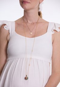 Des colliers en couches dorés avec un pendentif circulaire et un charme coloré. La robe est blanche avec des manches à volants et un tissu fluide.