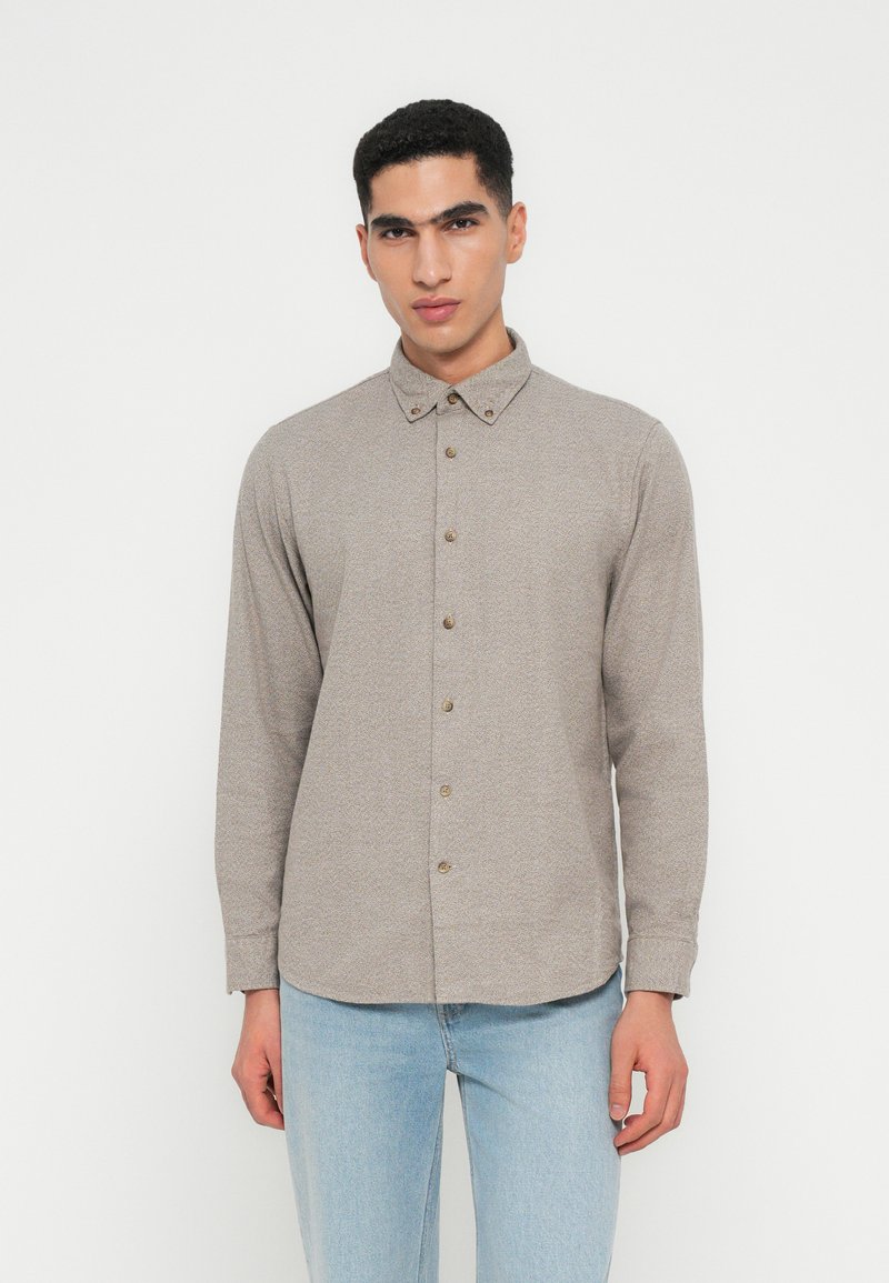 Giovane uomo che indossa una camicia button-down beige a maniche lunghe e jeans azzurri chiari, in piedi contro uno sfondo bianco semplice.