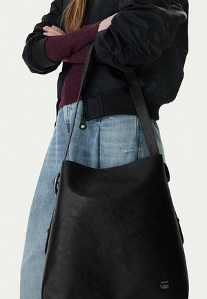 Person wearing black jacket and purple sweater carries a large black leather shoulder bag with long straps.