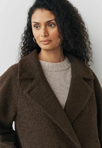 Manteau en laine texturé marron avec un col châle, présentant un look superposé sur un pull en tricot gris clair. Accents subtils et coupe oversize.