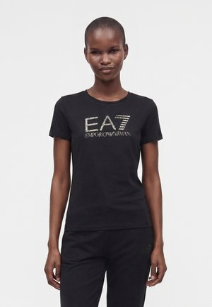 Naine kannab musta lühikeste varrukatega T-särki, millel on valge tekstiga "EA7 Emporio Armani" logo, sobitatud mustade pükstega, seisab valgel taustal.