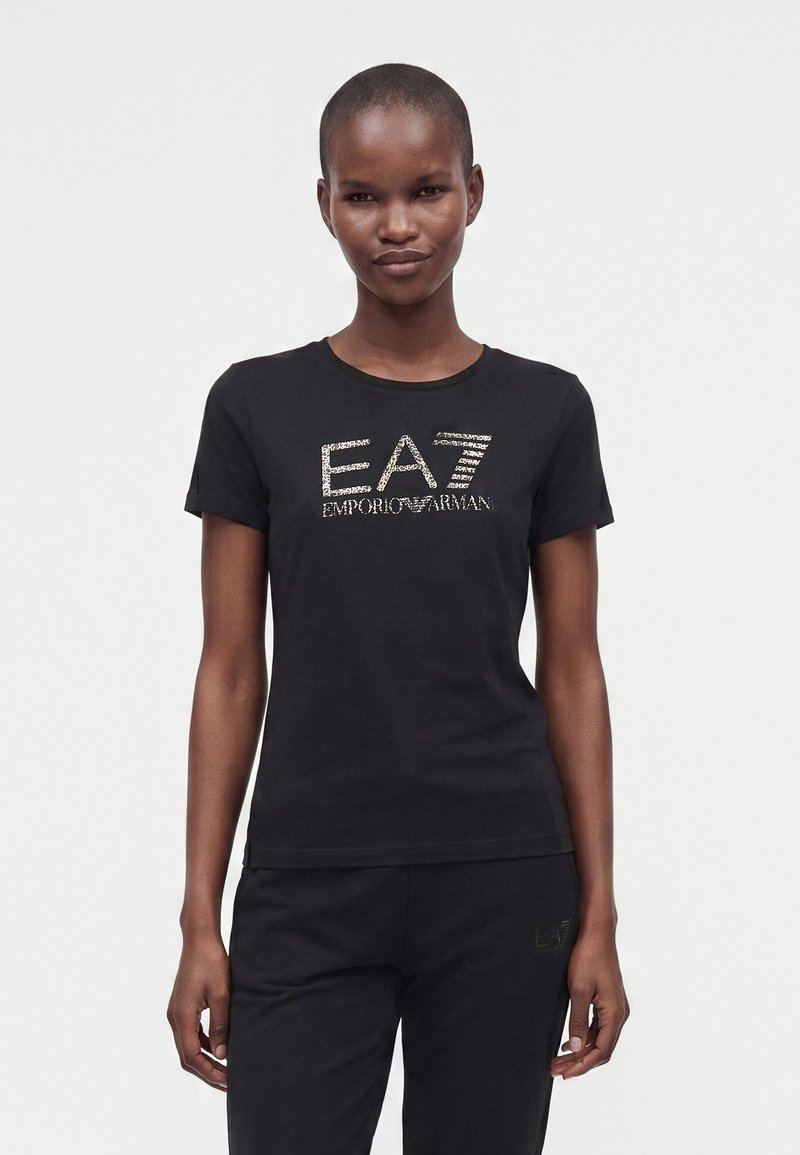 Γυναίκα που φοράει μαύρο κοντομάνικο T-shirt με το λογότυπο "EA7 Emporio Armani" σε λευκό κείμενο, σε συνδυασμό με μαύρο παντελόνι, στέκεται σε λευκό φόντο.