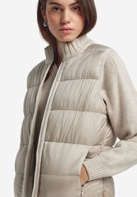 Fleecerad väst i beige, med hög ribbad krage, dragkedja och sidofickor. Slät, vadderad textur med horisontella sömmar.