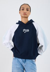 Hoodie blu navy con maniche raglan bianche e azzurro chiaro. Presenta una grafica con la scritta 'Paris'. Il tessuto sembra morbido e texturizzato.