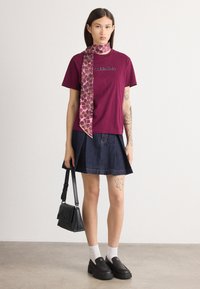 Burgunderfarget T-skjorte med Calvin Klein-logo, kombinert med et jeanstynne skjørt. Har et mønstrete rosa skjerf og svarte slip-on sko. Holder en svart håndveske.