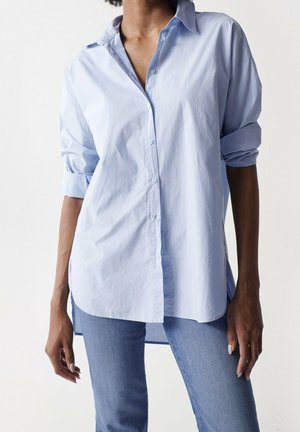 Personne portant une chemise bleu clair à boutons avec manches retroussées et un jean bleu, debout devant un fond blanc uni.