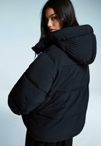 Zwarte pufferjack met een gequilte textuur, oversized silhouet en een grote capuchon. De stof lijkt soepel en lichtgewicht te zijn.