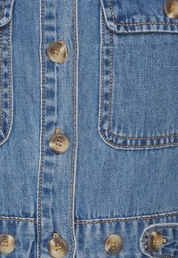 Jaqueta de jeans azul claro com costura visível, botões em tom dourado e dois bolsos no peito. Tecido texturizado com um design clássico.