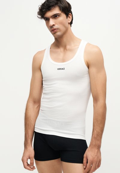 Jeune homme portant un débardeur blanc Versace et un boxer noir, debout devant un fond clair uni.