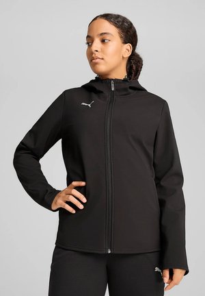 Femme aux cheveux foncés en chignon bas, portant une veste zippée noire Puma et un pantalon assorti, debout avec une main sur la hanche.