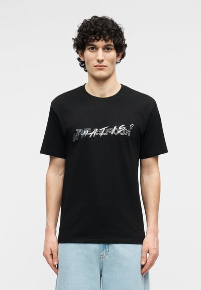 Νεαρός άνδρας με σγουρά μαλλιά, που φοράει μαύρο T-shirt με σχέδιο και ανοιχτού μπλε τζιν, στέκεται μπροστά σε σκονισμένο ανοιχτό γκρι φόντο.