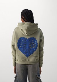 Felpa verde oliva con una grafica a forma di cuore con costole blu sul retro. Presenta un cappuccio con coulisse e polsini a coste. Il testo sotto la grafica recita "isola di cuori spezzati."