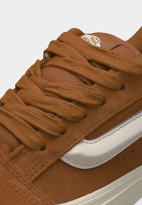 Baskets en daim marron avec des lacets épais marron, une bande latérale blanche et le logo Vans Off the Wall visible sur la languette.