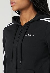 Femme portant un sweat à capuche zippé noir Adidas avec des bandes blanches sur les épaules et un logo Adidas sur la poitrine, gros plan sur le haut du torse.