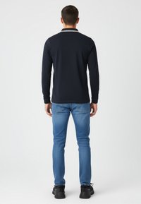 Marineblaue Langarm-Polo-Shirt mit schlichter Gestaltung, ausgestattet mit einem weißen Kragenbesatz, kombiniert mit hellblauen Jeans und schwarzen Sneakers.