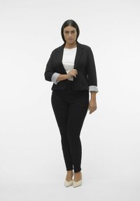 Sort blazer med stribede manchetter, hvid skjorte og sorte jeans. Modellen har beige stiletter med blokhæl på, stående mod en ensfarvet baggrund.