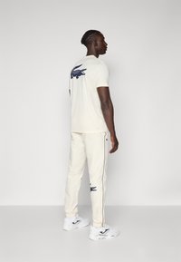 Maglietta a maniche corte color crema con logo di coccodrillo blu navy sulla schiena, abbinata a pantaloni color crema con dettagli blu navy e polsini elasticizzati, indossata con sneaker bianche.