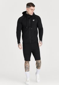 Musta vetoketjullinen huppari, jossa on nyörit ja logo rinnassa, yhdistettynä samansävyisiin mustiin shortseihin. Varustettu ribbineuleisine hihansuineen ja slim fit -leikkauksella.