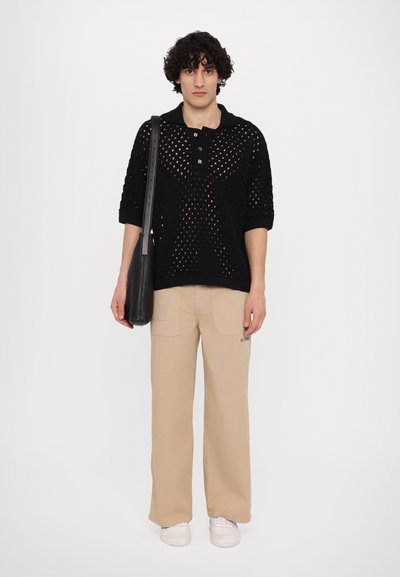 Polo en maille noire avec un motif en mesh, manches courtes et trois boutons, associé à un pantalon large beige et des baskets blanches. Sac à main noir.