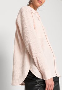Mujer que lleva una camisa de botón de gran tamaño en un suave tono de rosa con textura, con un bolsillo delantero y pantalones de cuero negros, vista de perfil con un pendiente de aro visible.
