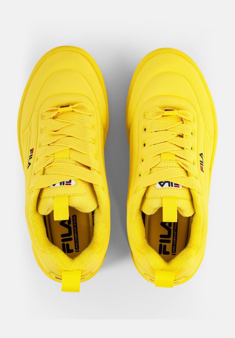 Zapatillas deportivas amarillas con una textura suave, cuello acolchado y cordones. Presentan un logo de la marca en el lateral y una lengüeta de talón reforzada.