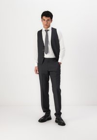 Tmavě šedý oblek na míru se vestou a kalhotami, doplněný o bílou košili a šedou kravatu. Černé kožené boty dotvářejí celý outfit.