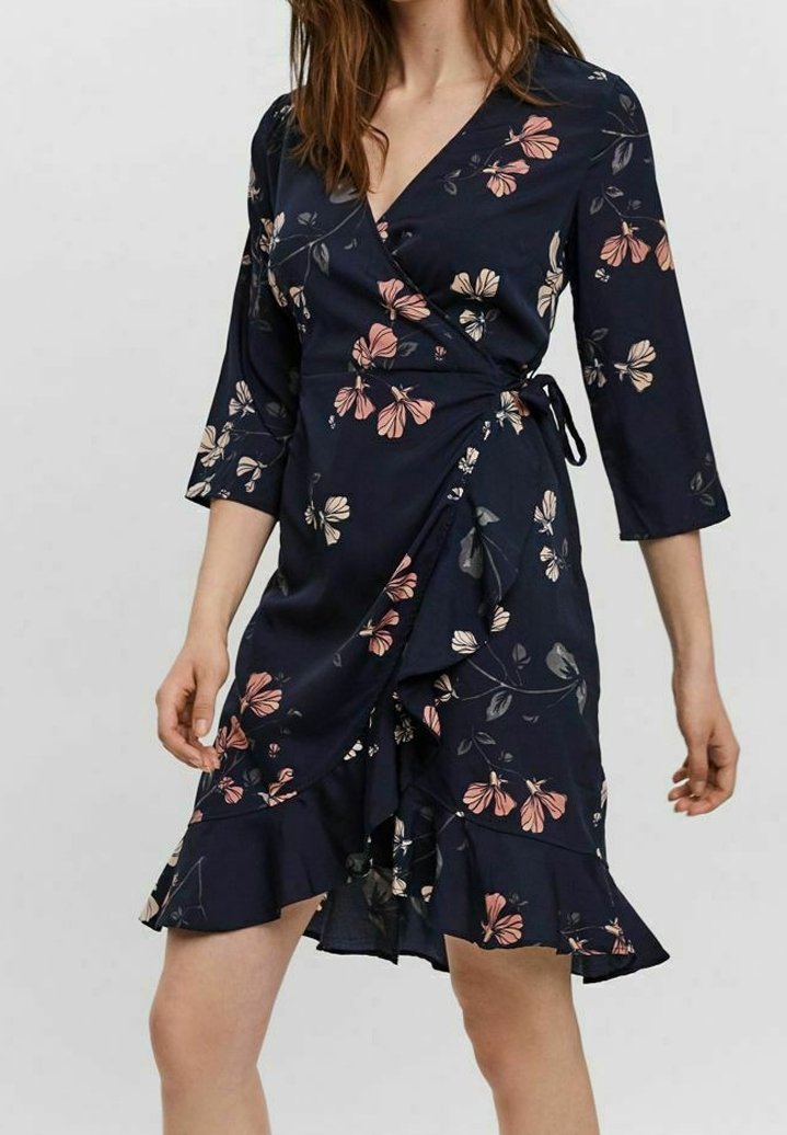 Femme portant une robe portefeuille bleu marine avec un imprimé floral rose et beige, manches trois-quarts et ourlet à volants.