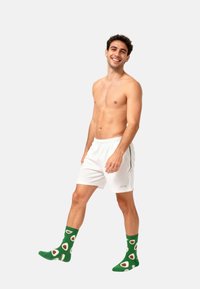 Smilende, brystløse mand går, iført hvide shorts og grønne sokker med avocado mønster imod en ensfarvet baggrund.