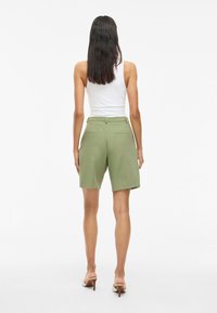 Pantalones cortos ajustados verdes con una textura suave, que cuentan con bolsillos laterales, una cinturilla con trabillas para cinturón y un corte relajado, combinados con una camiseta sin mangas blanca.