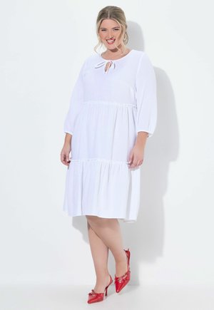 Lächelnde Frau, die ein weißes, langärmeliges, gestuftes Kleid mit vorderer Bindenausschnitt trägt, kombiniert mit roten spitzen Slingback-Absätzen.