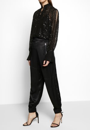 Kobieta ubrana w przezroczystą czarną bluzkę z kwiatowymi wzorami, czarne spodnie z wysokim stanem zapinane na boczne guziki oraz czarne sandały na wysokim obcasie.