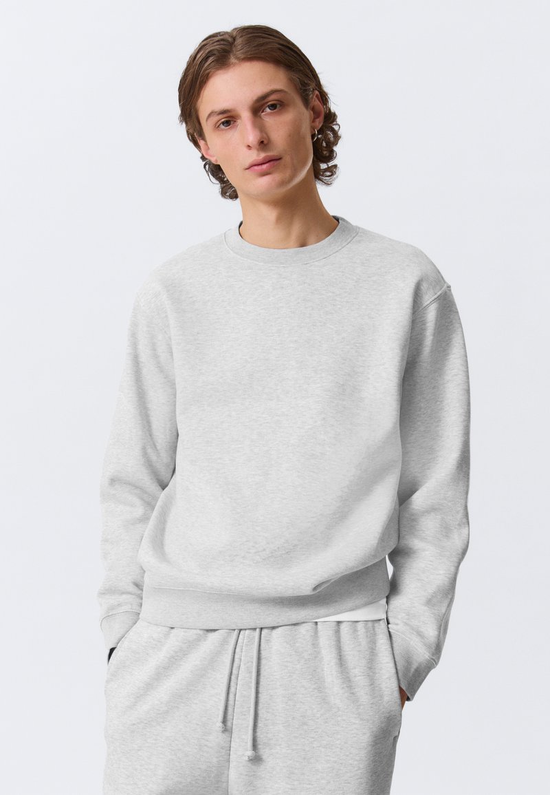 Svetlosiva majica dugih rukava od mekog materijala, s okruglim izrezom, dugim rukavima i rebrastim manžetnama i rubom. Uparena s odgovarajućim trenirkama.