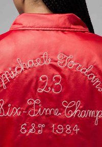 Punainen satiinitakki, jossa valkoisella kirjailtu teksti "Michael Jordan 23 Six-Time Champ EST 1984" eri kursiivikirjasintyyleillä.