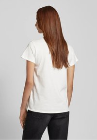 T-shirt bianco in cotone con maniche corte e scollatura rotonda. Design semplice, mostrata di spalle, abbinata a un paio di jeans neri.