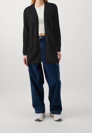 Schwarze gestrickte Cardigan mit offenem Vorderteil, langen Ärmeln und zwei vorderen Taschen, kombiniert mit lässigen blauen Jeans und weißen Sneakers.