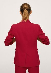 Blazer rosso aderente con un design strutturato, apertura posteriore centrale e maniche arrotolate. Texture liscia con dettagli hardware minimi.