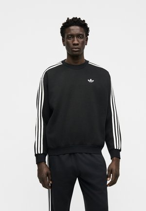 Uomo con capelli corti e ricci, indossa una felpa nera Adidas e pantaloni con dettagli bianchi a tre strisce, in piedi davanti a uno sfondo chiaro uniforme.