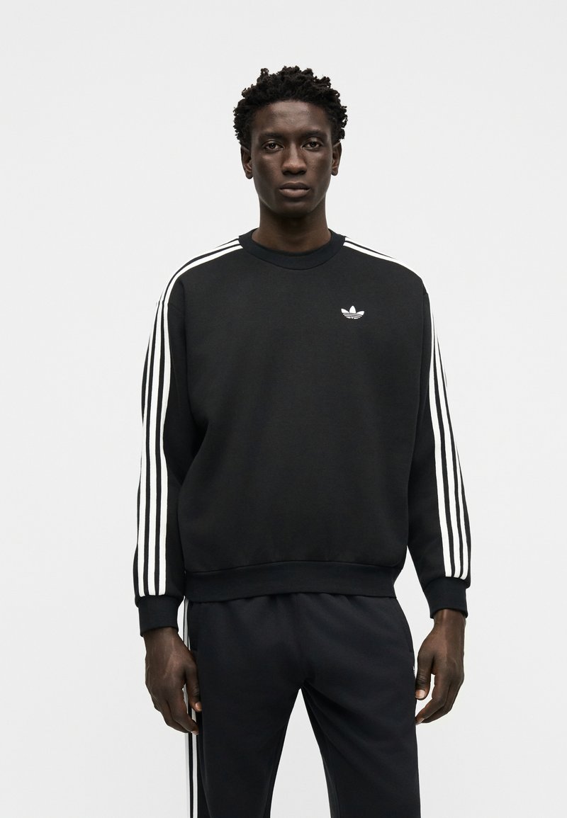 Man with short curly hair wearing black Adidas sweatshirt and pants with white three-stripe details, standing against a plain light background.