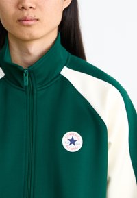 Πράσινο φούτερ με φερμουάρ και κρεμ λεπτομέρειες, που φέρει το στρογγυλό λογότυπο Converse All Star στο στήθος, διαθέτοντας ψηλό γιακά και λείες υφές.