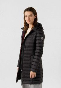 Mujer con una chaqueta acolchada negra hasta la rodilla y capucha, de pie frente a un fondo claro y liso, mirando ligeramente hacia la izquierda.