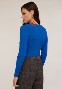 Donna con lunghi capelli castani, indossa un top a maniche lunghe blu a coste, orecchini a cerchio dorati e pantaloni grigi a quadri a vita alta con cintura marrone, rivolta a sinistra.