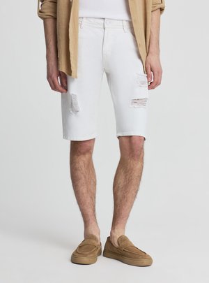 Mężczyzna w białych podniszczonych krótkich spodenkach jeansowych, beżowych mokasynach z zamszu i brązowej podwiniętej koszuli na białym topie, stojący na jednolitym tle.