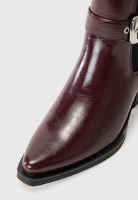 Stivale alla caviglia bordeaux realizzato in materiale sintetico lucido, con punta affusolata, elastico nero laterale e un'accents di fibbia metallica.
