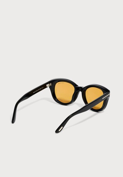 Lunettes de soleil en acétate noir avec des montures épaisses et des verres ronds teintés ambre, ouvertes et posées sur une surface blanche.